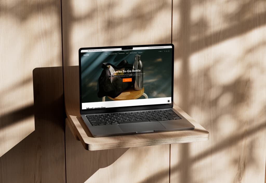 Laptop on a light wood shelf showing the Aarke To-Go Bottle campaign and an orange Shop now button