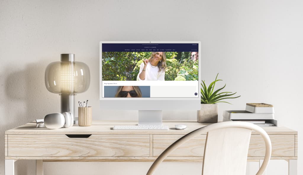 iMac on a light wood desk showing the Sidney Garber jewelry website with a navy SIDNEY GARBER header, a hero of a woman in a white knit sweater outdoors, and Shop Signature Pieces below